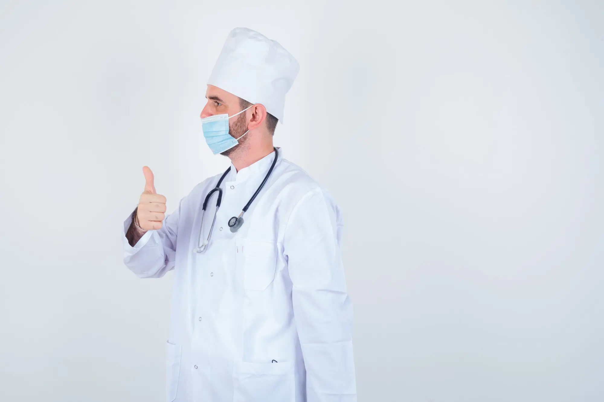 Gutaussehender Mann mit um den Hals hängendem Stethoskop, der den Daumen nach oben zeigt, in weißem medizinischen Laborkittel und mit Maske, blickt selbstbewusst, Frontalansicht, praxisnah und nachvollziehbar.